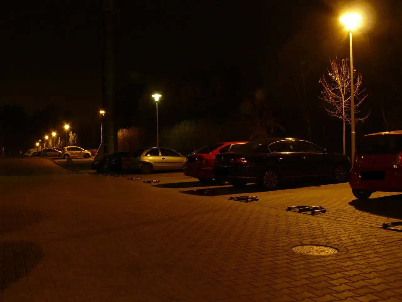 Nocny parking oświetlony lampami sodowymi, tworzącymi ciepłą, żółtą poświatę. W tle widać rząd samochodów.