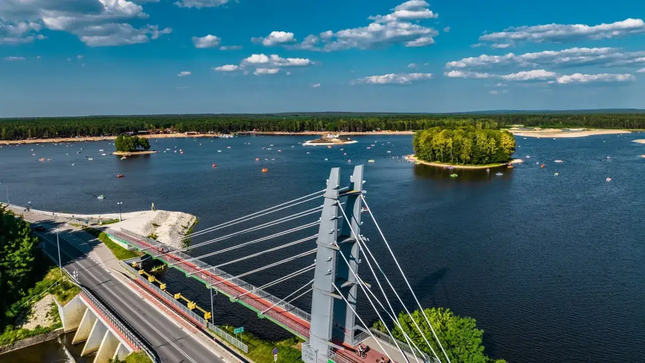 Sielpia jezioro tętni życiem. Wiele łódek i kajaków pływa po wodzie, a na brzegu widać plażowiczów. Most nad jeziorem dodaje uroku.