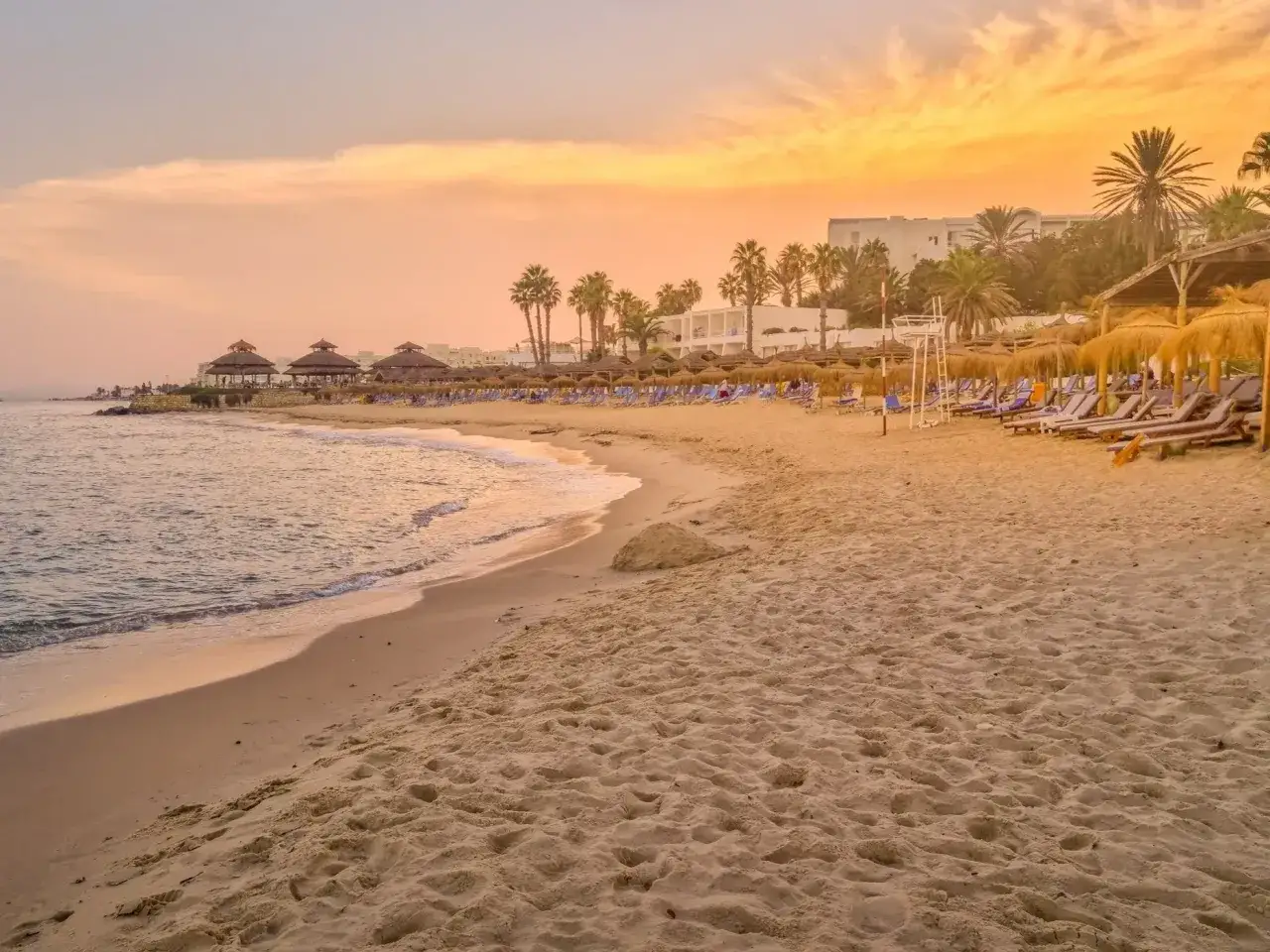 Piękna plaża w Tunezji w lipcu. Słońce zachodzi, tworząc złote niebo nad palmami i leżakami. Idealna pogoda na wakacje.