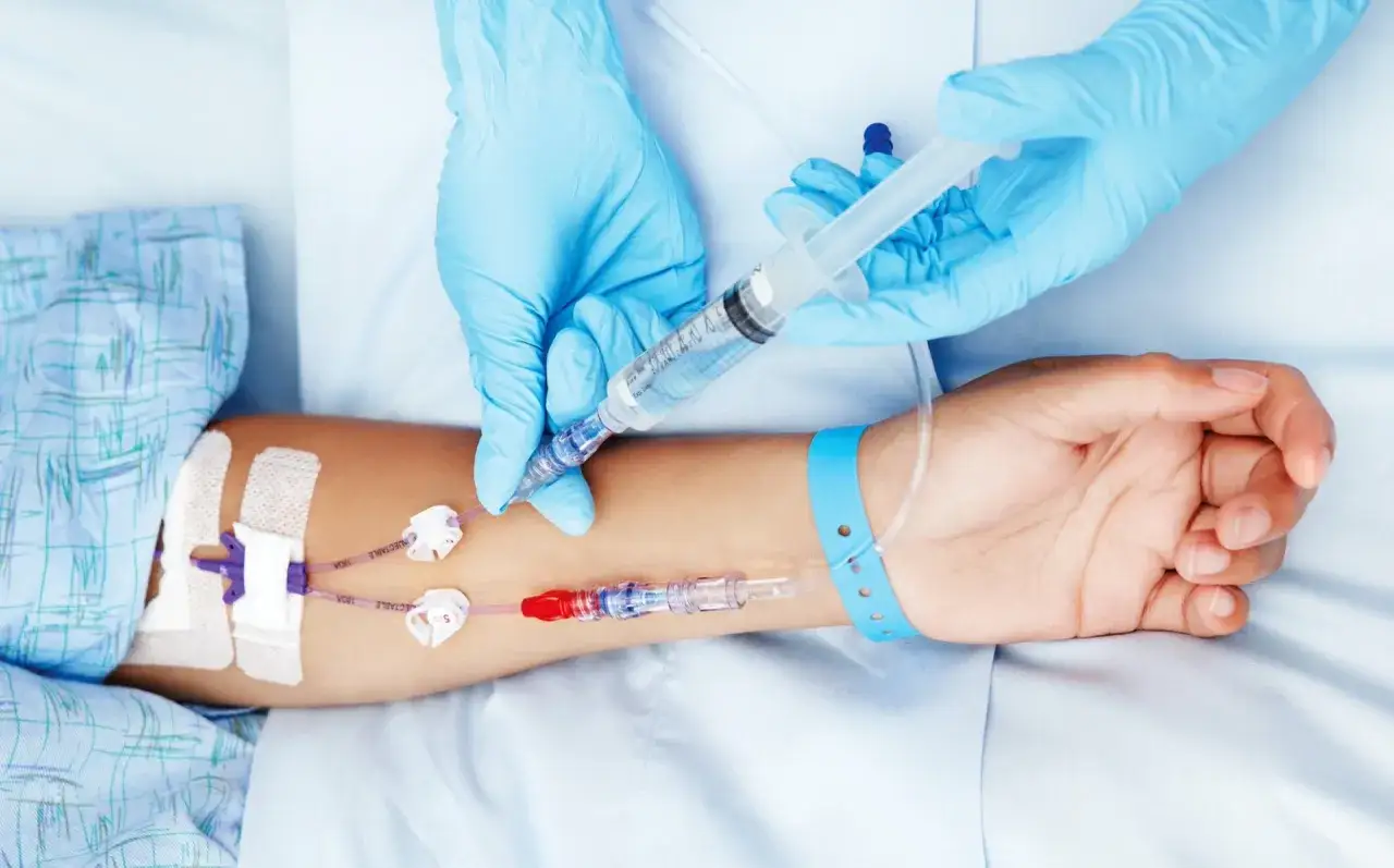 Mani guantate in medicina preparano un'iniezione. Un braccio con catetere venoso centrale riceve un farmaco.