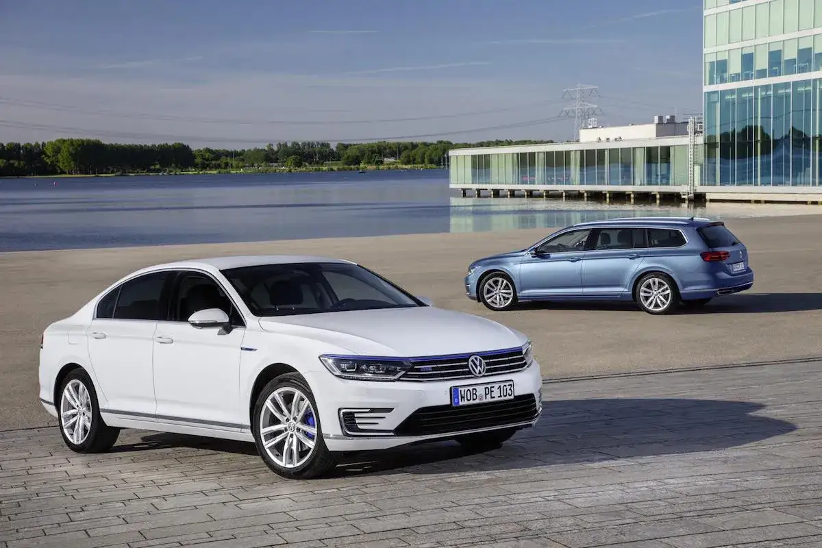 Dos coches Volkswagen, uno blanco sedán y otro azul familiar, estacionados cerca de un lago y un edificio moderno. ¿Cuál es el permiso de coche que necesitas para conducirlos?