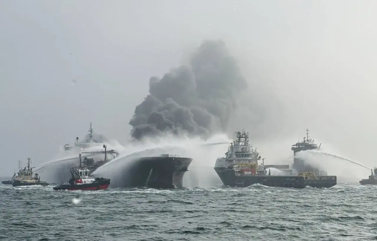 Plusieurs remorqueurs luttent contre un incendie sur un navire, projetant de l'eau sur la structure en feu. La scène évoque une **collision bateau** dramatique.