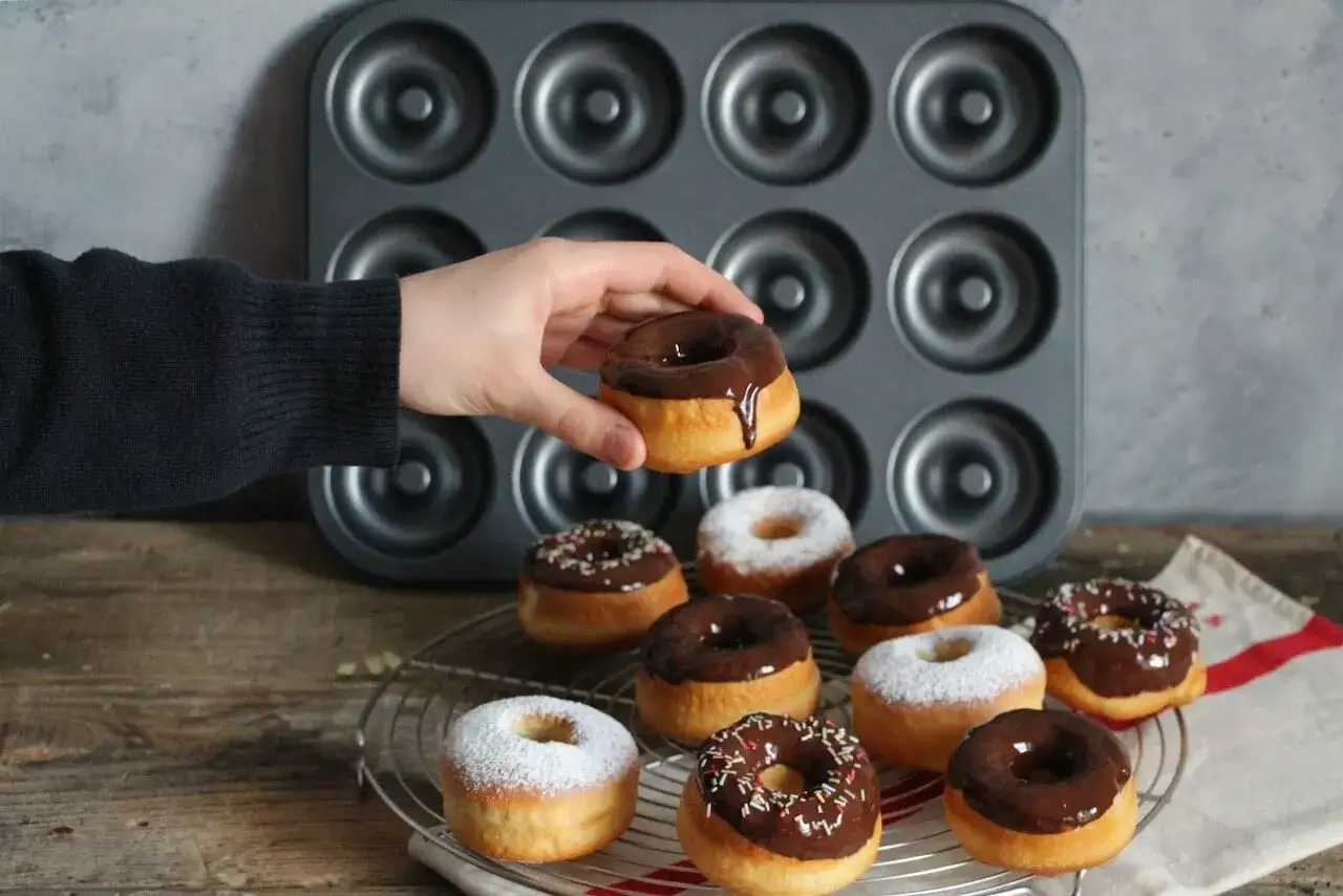 Une main tient un donut glac&eacute; au chocolat, pr&ecirc;t &agrave; &ecirc;tre d&eacute;gust&eacute;. Une belle collection de donuts au four, certains saupoudr&eacute;s de sucre glace, d'autres de gla&ccedil;age au chocolat et de vermicelles color&eacute;s, attendent sur une grille.