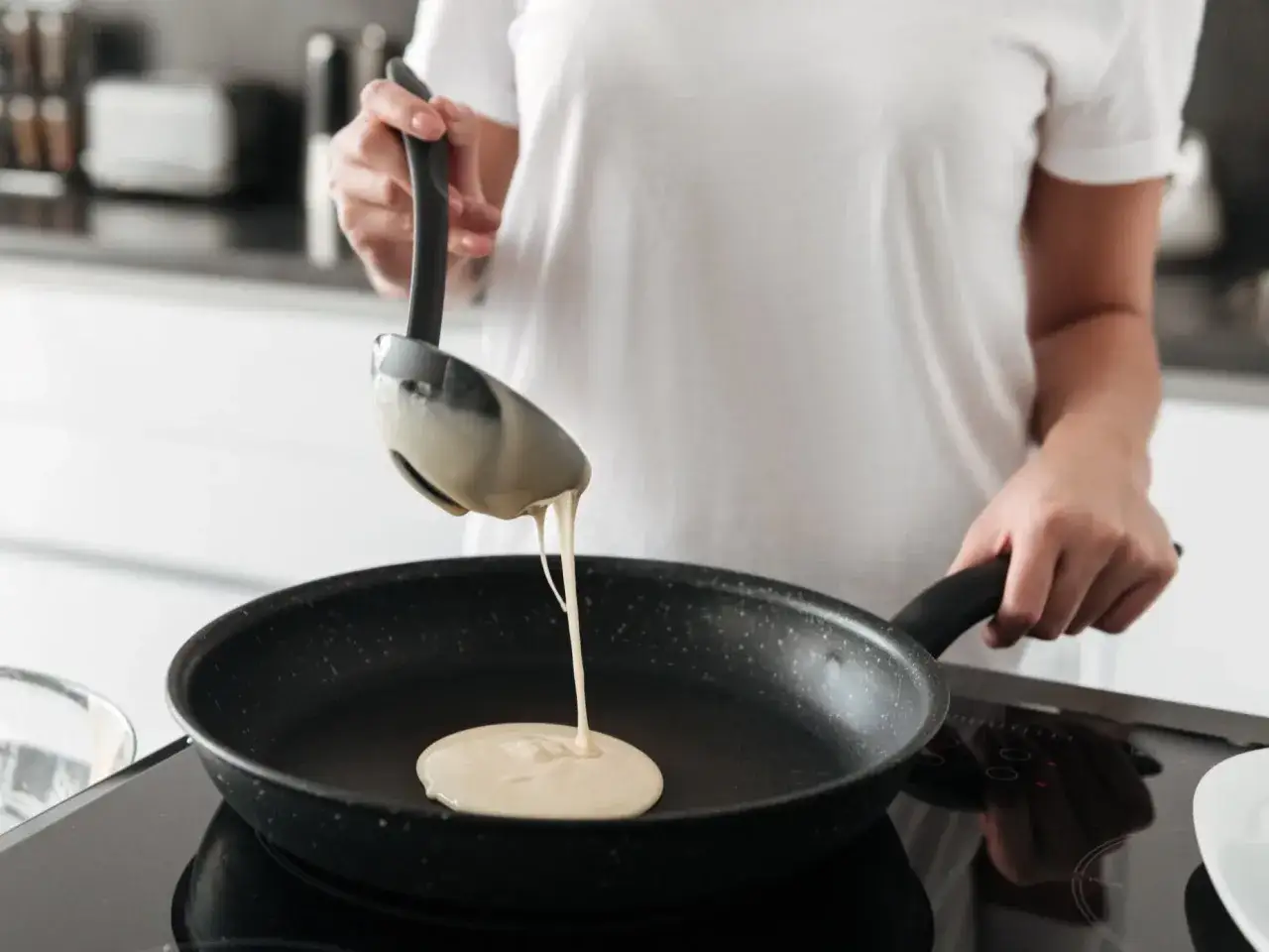Osoba w białej koszulce nalewa ciasto na patelnię, przygotowując pyszne naleśniki bez jajek.