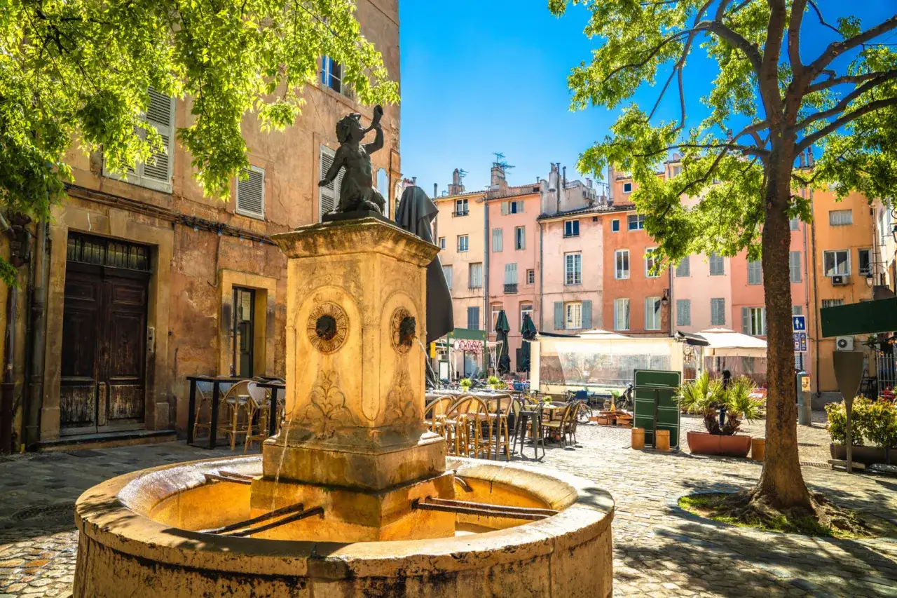 Ein Brunnen mit einer Statue in Aix-en-Provence, umgeben von bunten Häusern und schattenspendenden Bäumen. Entdecken Sie die Aix-en-Provence Geheimtipps!