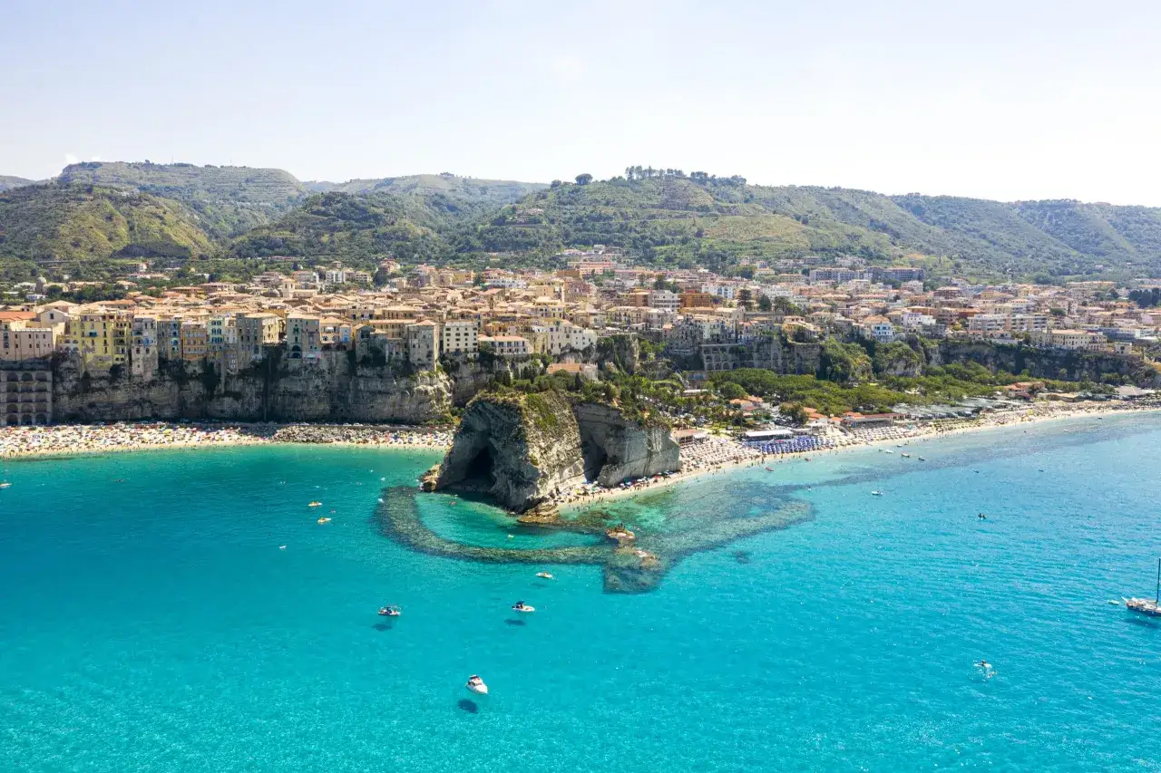Vue aérienne de Tropea, Calabre, avec ses maisons colorées perchées sur la falaise, une plage bondée et des eaux turquoise cristallines.