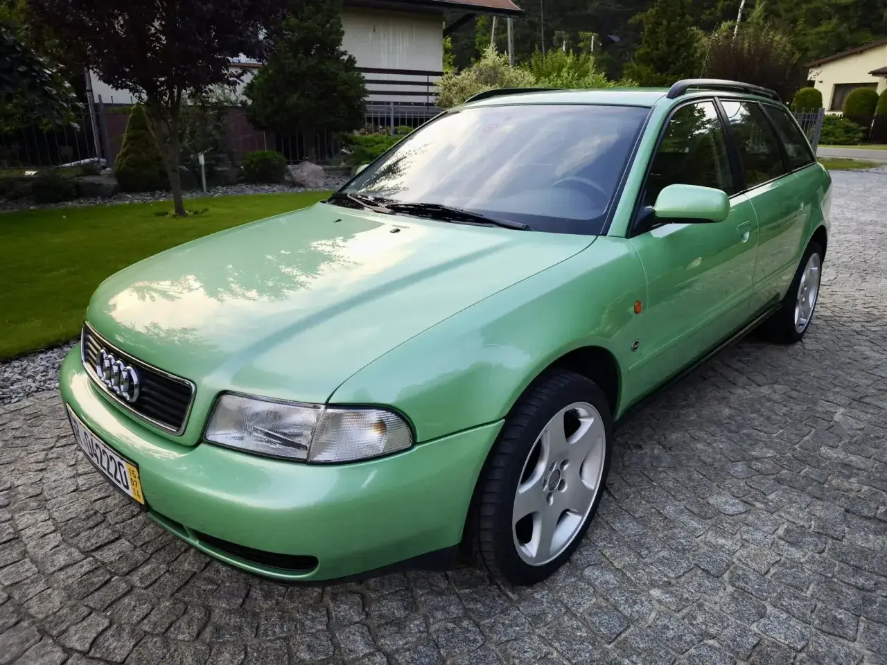 Zielone Audi B5 Avant na brukowanym podjeździe, otoczone zadbanym ogrodem i domem.