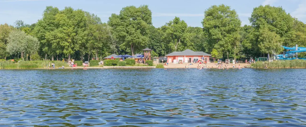 Sommerliche Szene an einem der **Badeseen Berlin** mit vielen Menschen am Strand und im Wasser, umgeben von grünen Bäumen.