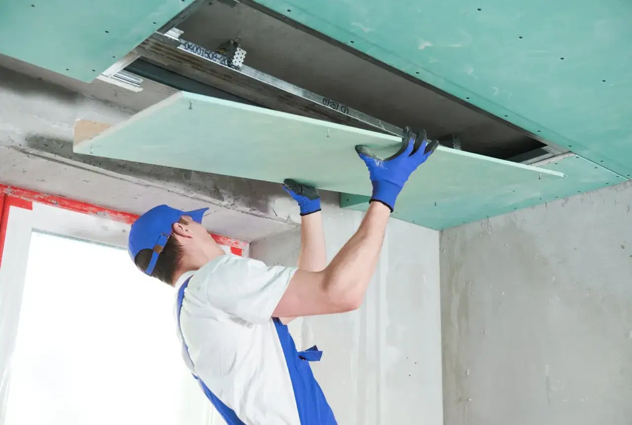 Pracownik montuje zieloną płytę gk na suficie podwieszanym. Standardowe wymiary płyty gk ułatwiają pracę.