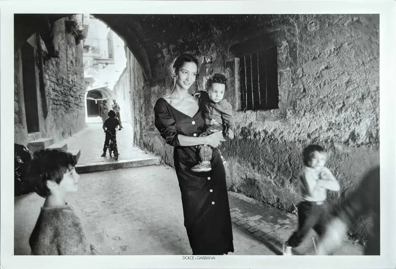 Una donna elegante tiene un bambino in un vicolo. Un bambino su una bici e un altro di spalle completano la scena, catturata dal **scianna fotografo**.