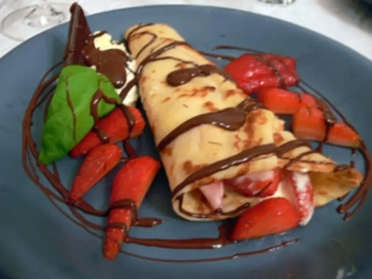Palatschinken mit Eis, Erdbeeren und Schokoladensauce auf einem blauen Teller.