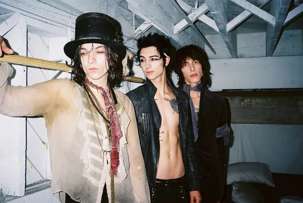Trzech członk&oacute;w Palaye Royale w stylowych, mrocznych strojach, z kapeluszem i sk&oacute;rzaną kurtką.
