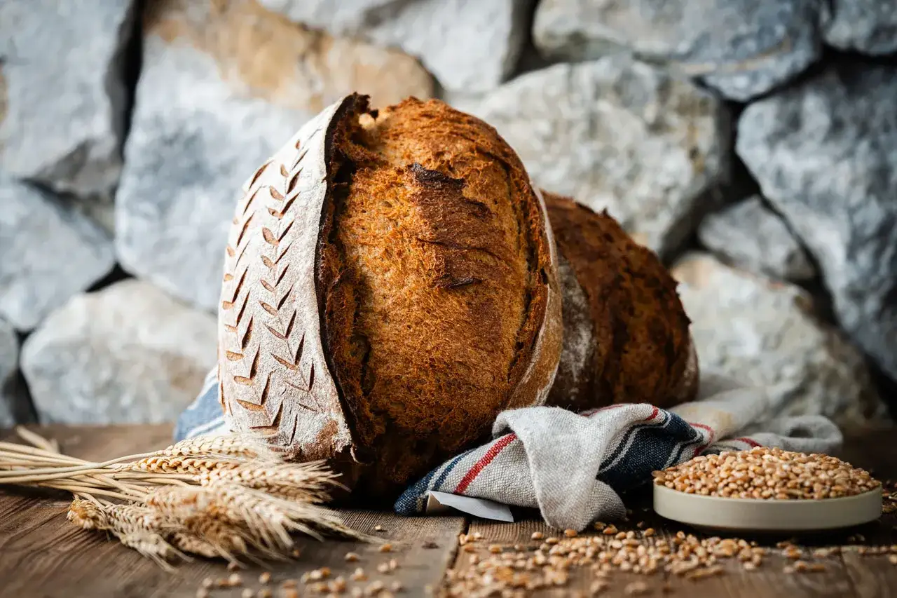 Zwei rustikale Brote, eines mit eingekerbtem Muster, liegen auf einem Holztisch. Weizen&auml;hren und eine Schale voller K&ouml;rner deuten auf ein k&ouml;stliches Ruchbrot Rezept hin.
