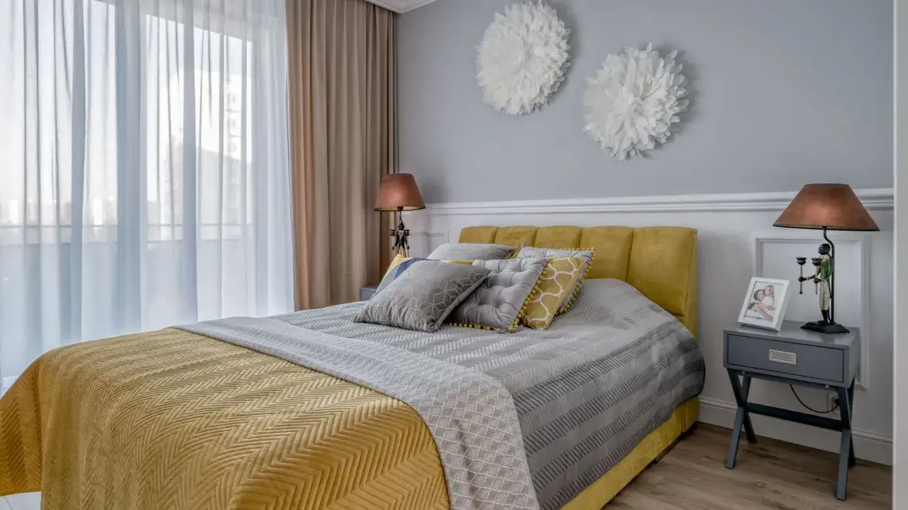 Żółty to świetny **jaki kolor do sypialni**! Wygodne łóżko z żółtym zagłówkiem, szare i złote poduszki, zasłony i lampki tworzą przytulną atmosferę.