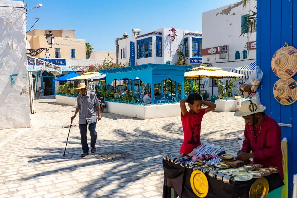 Djerba miasto: starszy mężczyzna z laską idzie brukowaną uliczką, obok stragan z pamiątkami i chłopiec w czerwonej koszulce.