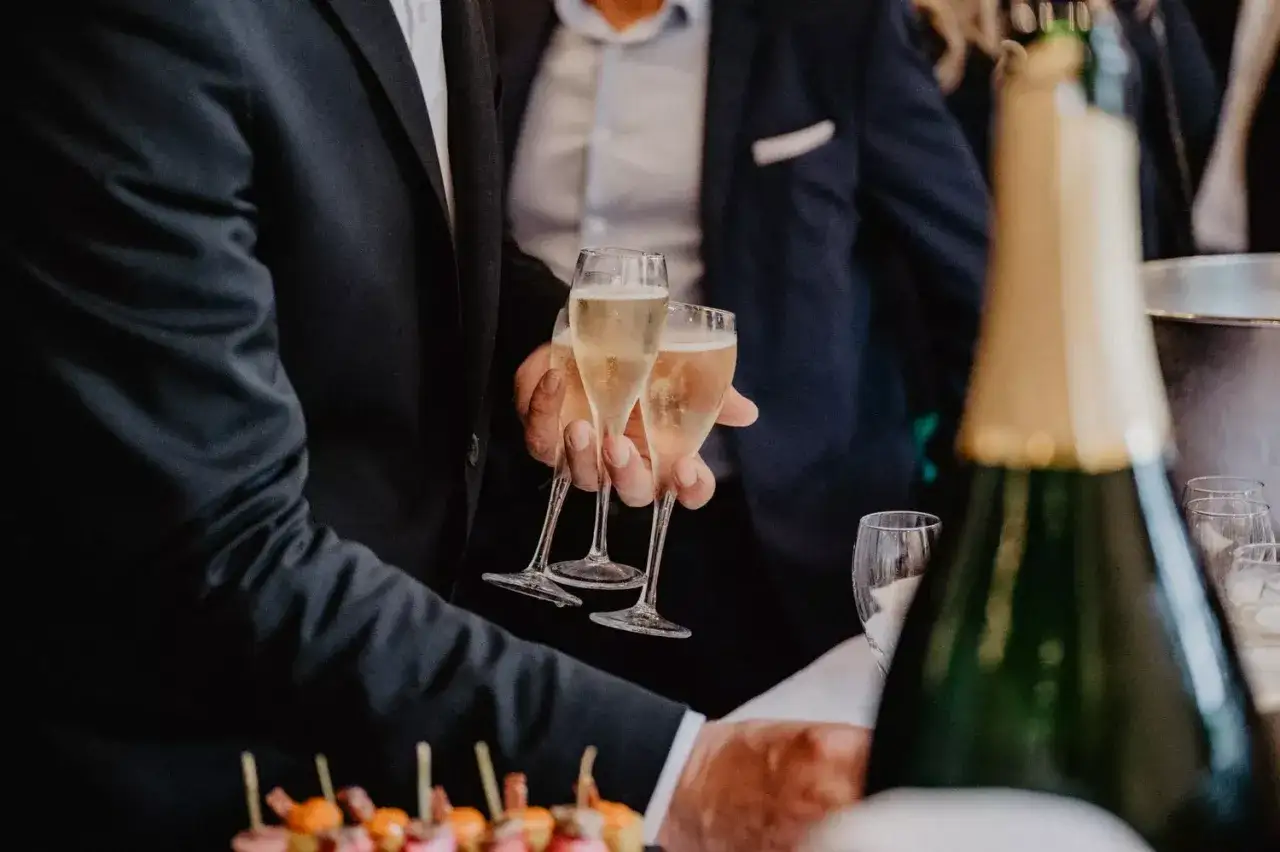 Des verres de champagne sont tenus par des mains gantées, prêts à être servis. La quantité de boisson pour ce mariage semble généreuse, avec une bouteille en arrière-plan.