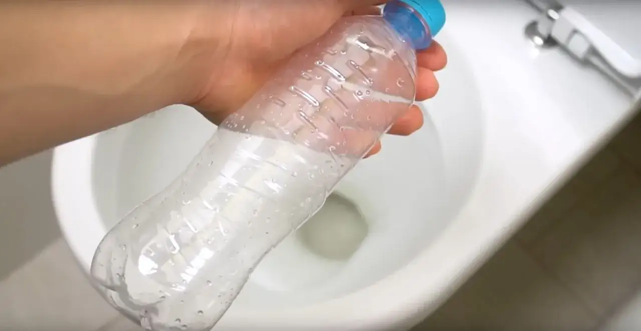 Dłoń trzyma plastikową butelkę z wodą nad białą muszlą klozetową. Dowiedz się, jak usunąć zapach z plastikowej butelki.