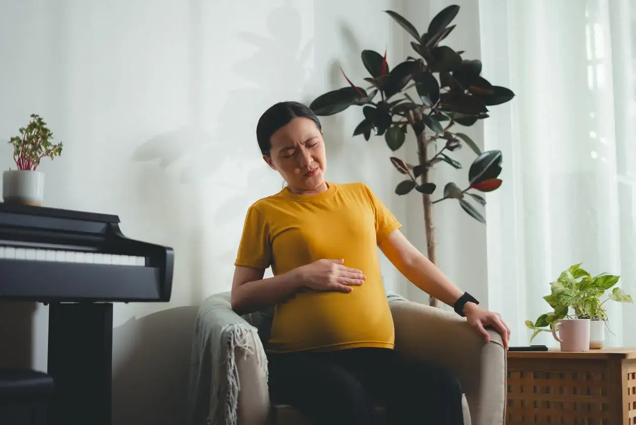 Kobieta w ciąży siedzi na fotelu, trzymając się za brzuch i wyrażając dyskomfort. Ból żeber w ciąży utrudnia jej relaks.