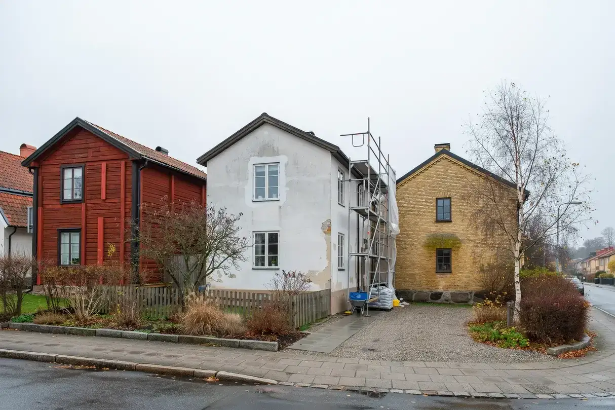 Hus med st&auml;llningar f&ouml;r att byta fasad. Ett r&ouml;tt tr&auml;hus, ett vitt hus under renovering och ett gult tegelhus.