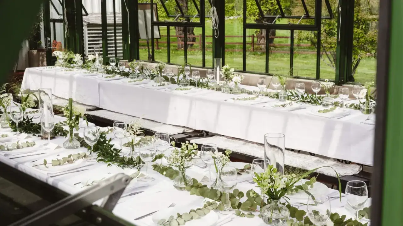 En vacker dukning i ett växthus med långbord, vita linnedukar, gröna blad och små blommor i glasvaser.