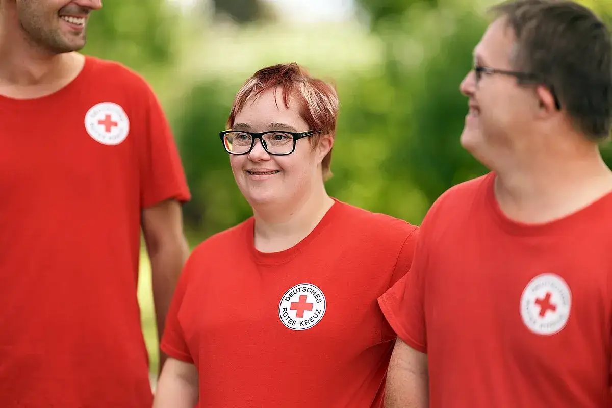 Drei Menschen im roten T-Shirt des Deutschen Roten Kreuzes. Eine Frau mit Down-Syndrom l&auml;chelt. Solche behinderungen sind Teil der Vielfalt.
