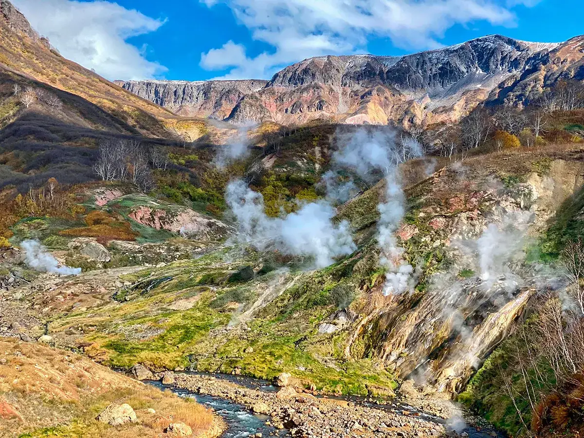 Dampf steigt aus einer geothermalen Landschaft in Russland auf, mit bunten Hügeln und einem Fluss.