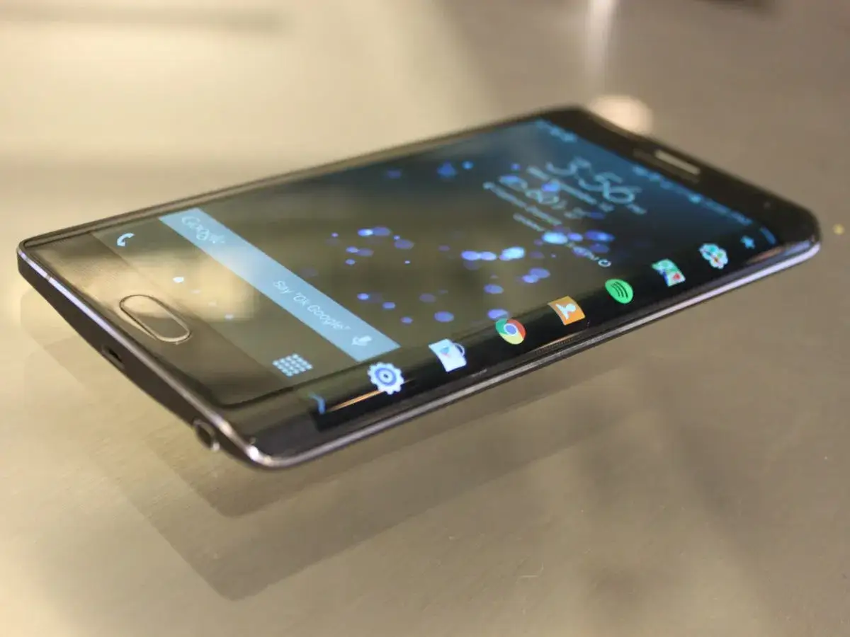 Nowoczesny **Samsung z zakrzywionym ekranem** leży na metalowej powierzchni. Na ekranie widać ikonki aplikacji i pasek wyszukiwania Google.