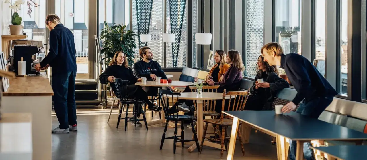 M&auml;nniskor fikar och samtalar vid bord i ett ljust, modernt caf&eacute;. En man g&ouml;r kaffe, ett exempel p&aring; multitasking svenska.