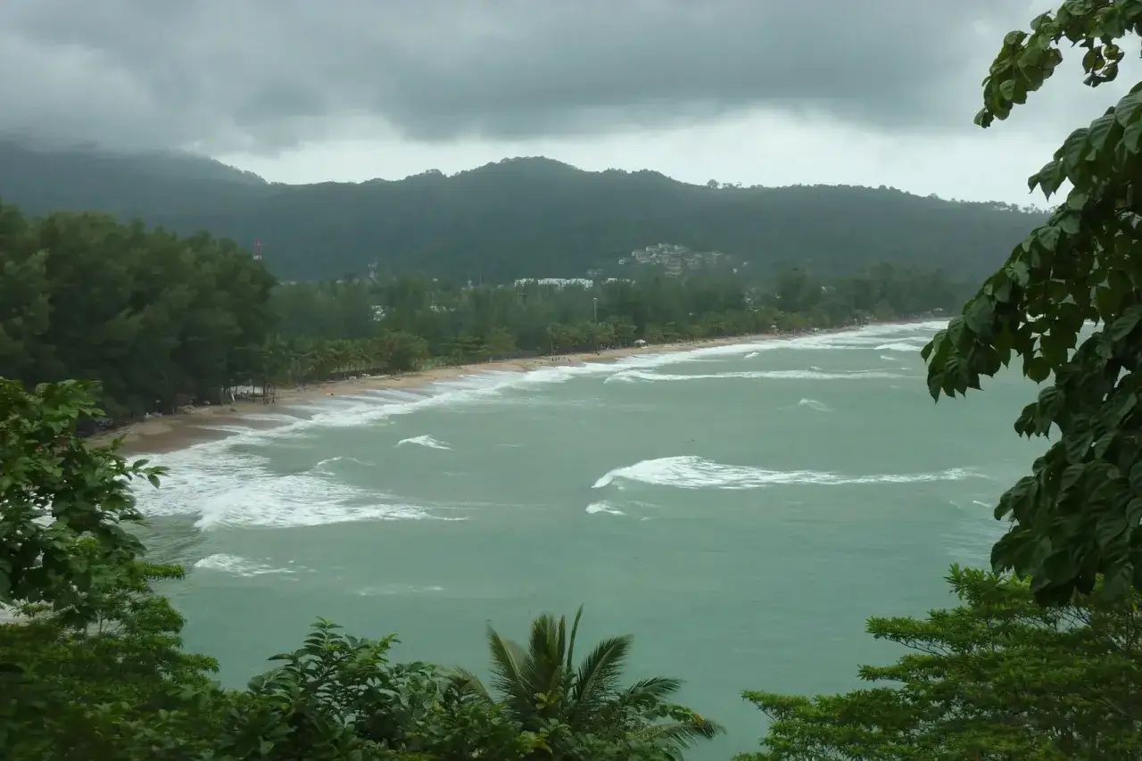 Khao Lak Regenzeit: Grauer Himmel über einem stürmischen Meer mit Wellen, die auf den Strand treffen. Palmen und üppige Vegetation säumen die Küste.