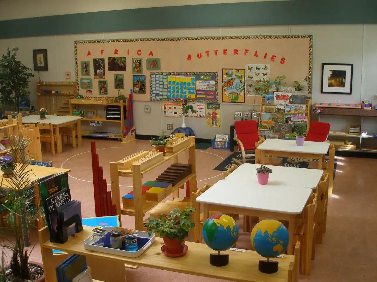 Klasa przygotowana zgodnie z zasadami metody Montessori. Widać globusy, materiały edukacyjne i tablicę z motywem Afryki i motyli.