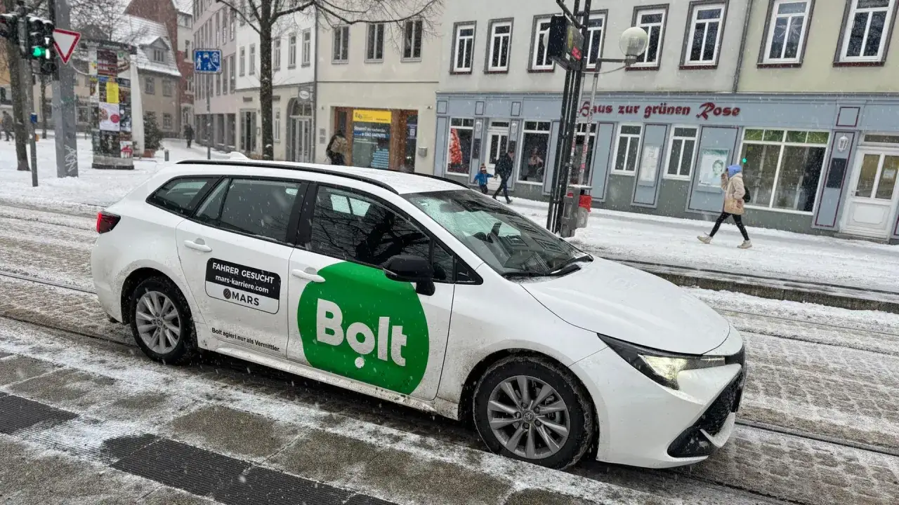 Ein wei&szlig;er Bolt-Taxi mit gr&uuml;ner Aufschrift steht im Schnee. Ein Schild sucht Fahrer.