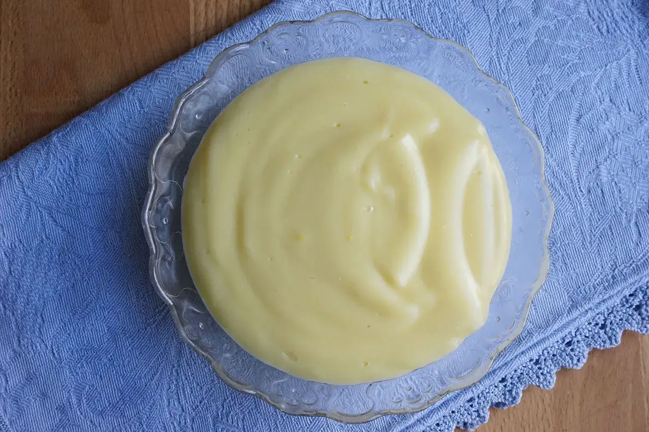 Crema pasticcera bimby, liscia e vellutata, servita in una ciotola di vetro decorata su un tovagliolo blu.