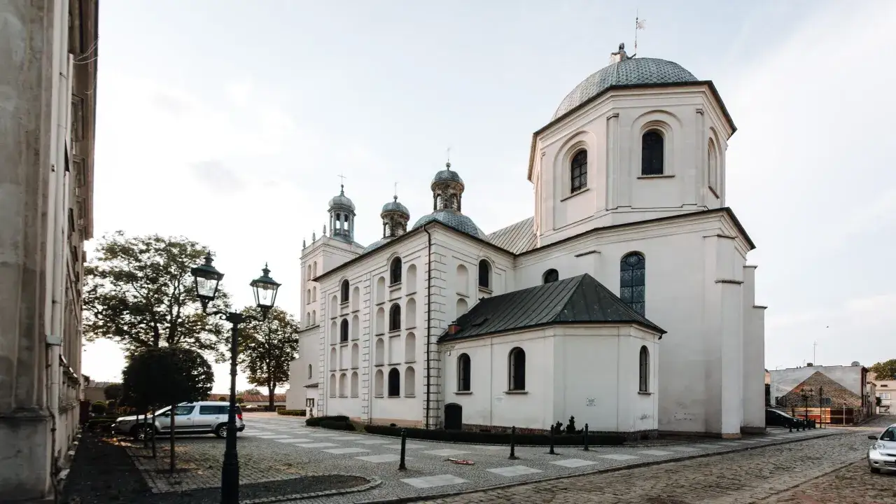 Majestatyczna fara w Grodzisku Wlkp. z charakterystycznymi kopułami i wieżami, otoczona brukowanym placem i zielenią.