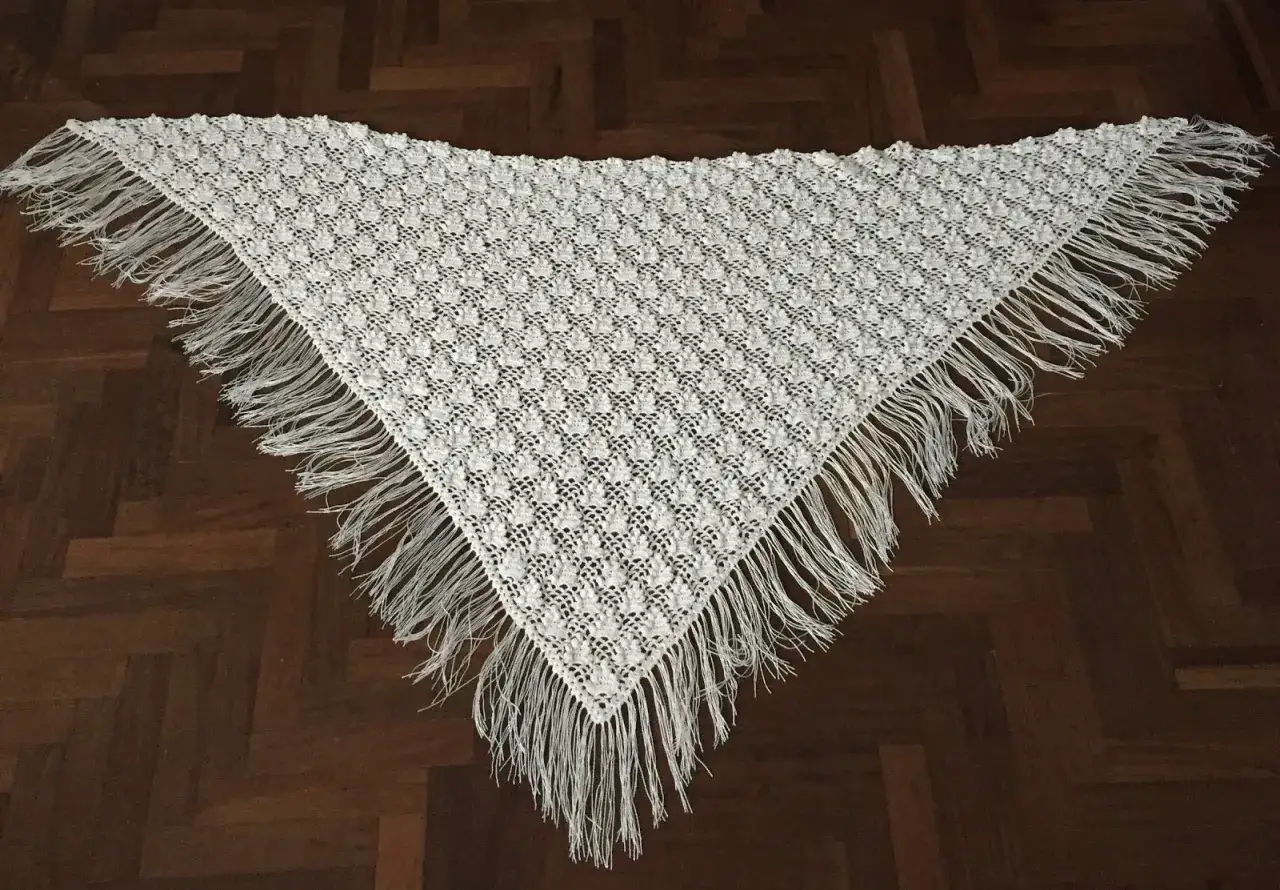 Un delicado pa&ntilde;uelo triangular crochet blanco, con un intrincado patr&oacute;n de encaje y flecos largos, descansa sobre un suelo de madera oscura.