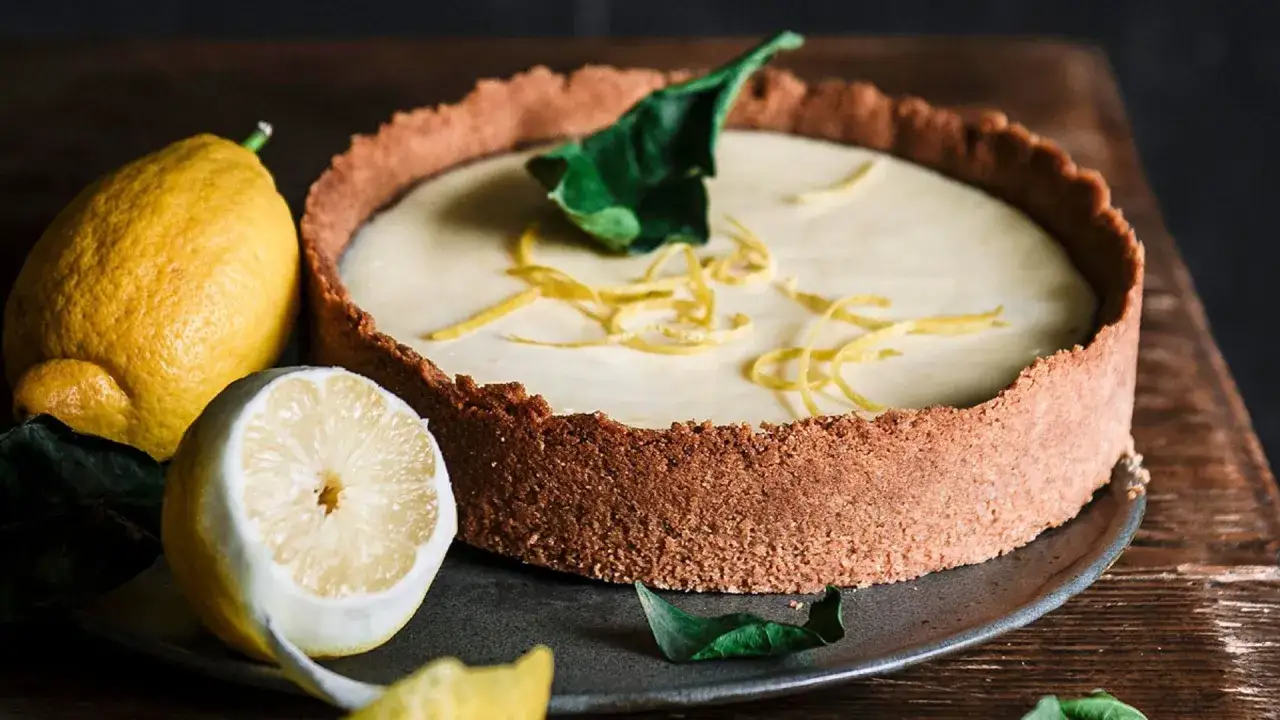Tarta de lim&oacute;n cremosa con base crujiente, decorada con ralladura y hojas verdes. Limones frescos acompa&ntilde;an este postre.