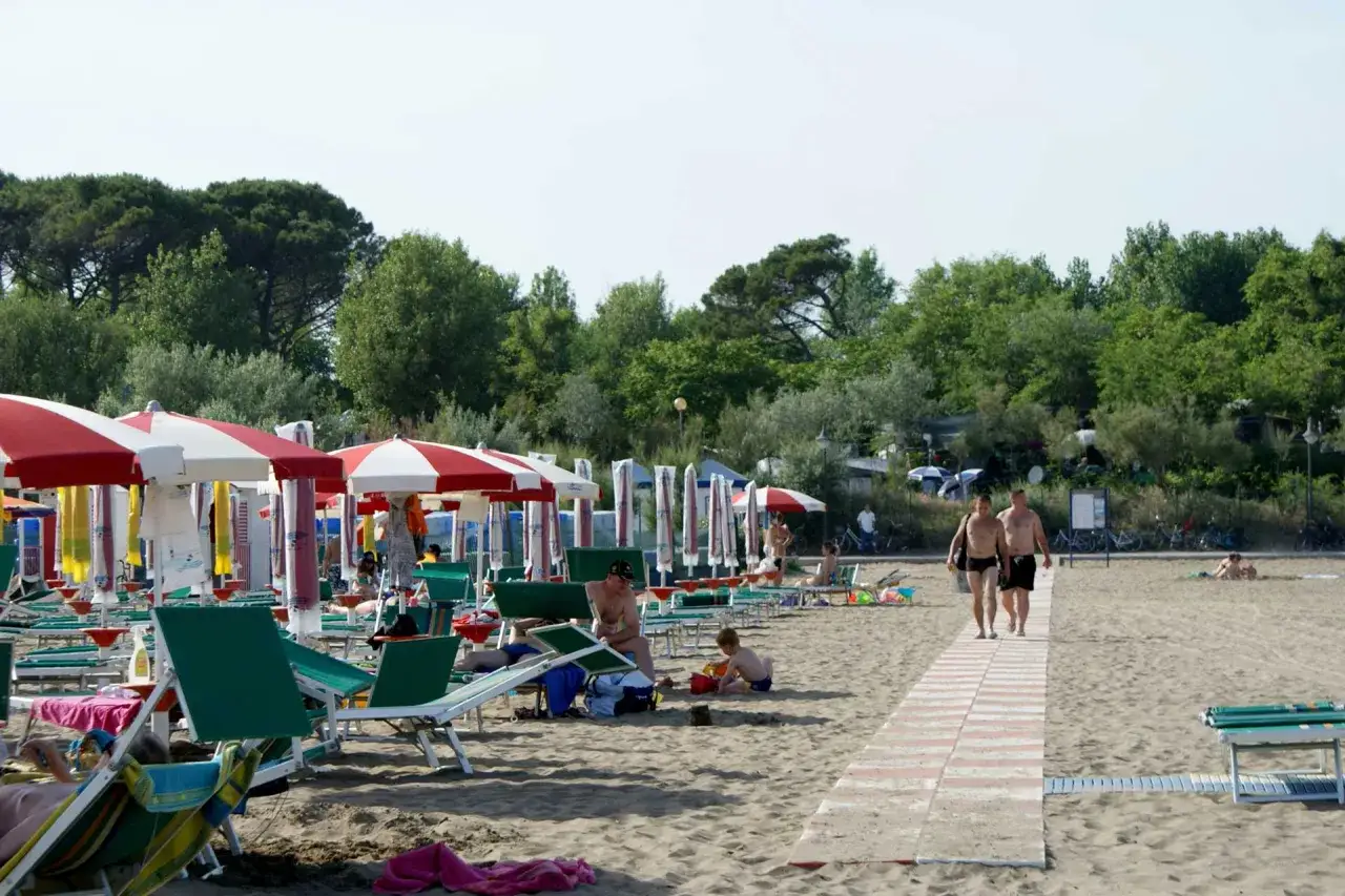 Plaża w Caorle, Santa Margherita. Rzędy parasoli i leżaków, ludzie odpoczywają, spacerują. W tle drzewa i camping.