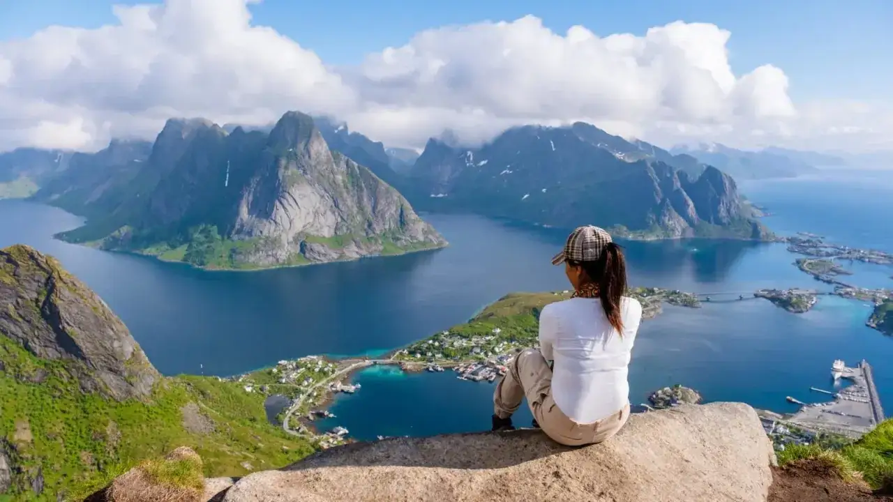 En person sitter på en klippa och blickar ut över en dramatisk kustlinje med berg och en by. En perfekt vy efter en härlig lofoten vandring.