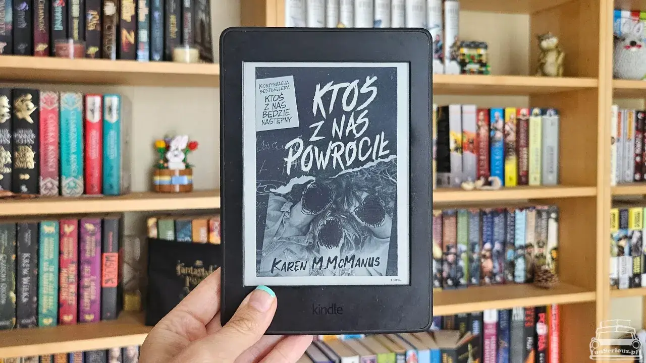 Ktoś z nas kłamie, a teraz ktoś z nas powrócił. Kindle z okładką książki na tle regałów pełnych książek.