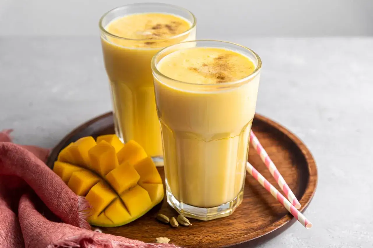 Två glas med krämig indisk yoghurtdryck, garnerad med kanel. Bredvid en skuren mango och kardemummakapslar på ett träfat.