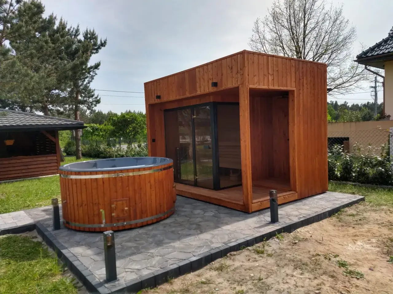 Drewniana balia z wodą, czyli co to jest jacuzzi, stoi obok nowoczesnej sauny w ogrodzie.