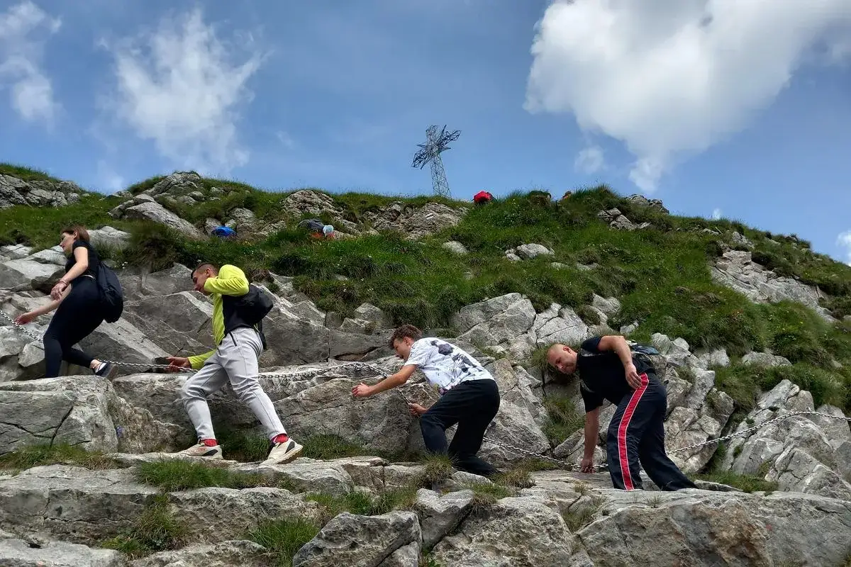 Turyści pokonują skalisty szlak na Giewont, używając łańcuchów. W tle widoczny krzyż.
