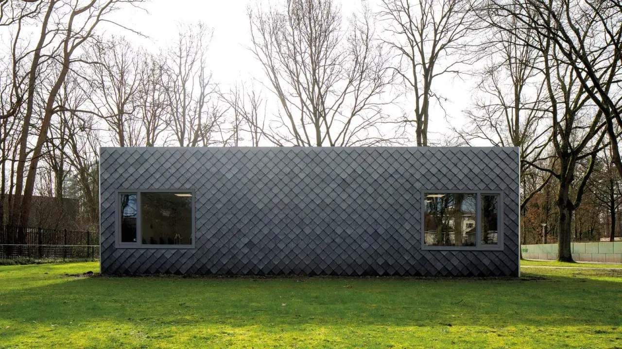 Modern building clad in dark, diamond-shaped recycled shingles, set against a backdrop of bare trees and green grass.