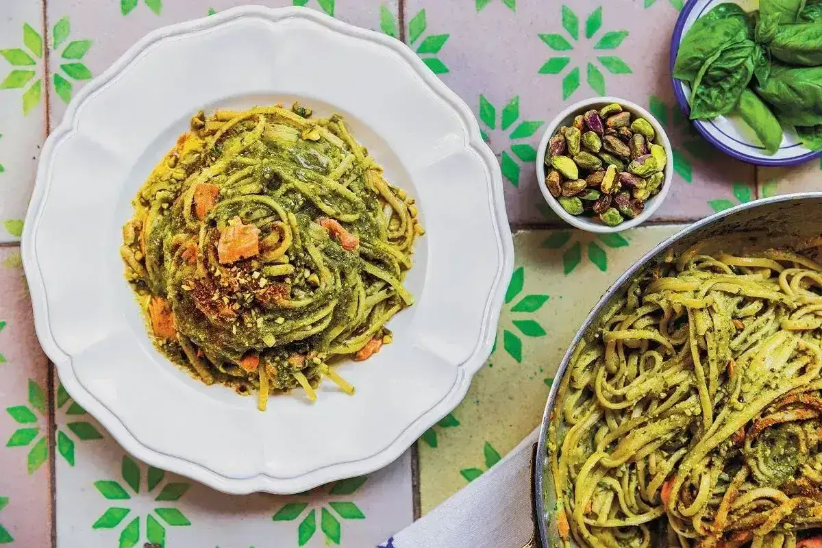 Makaron z pesto ze słoika, posypany pistacjami i kawałkami suszonych pomidorów, podany na białym talerzu. Obok miseczka z pistacjami i listkami bazylii.