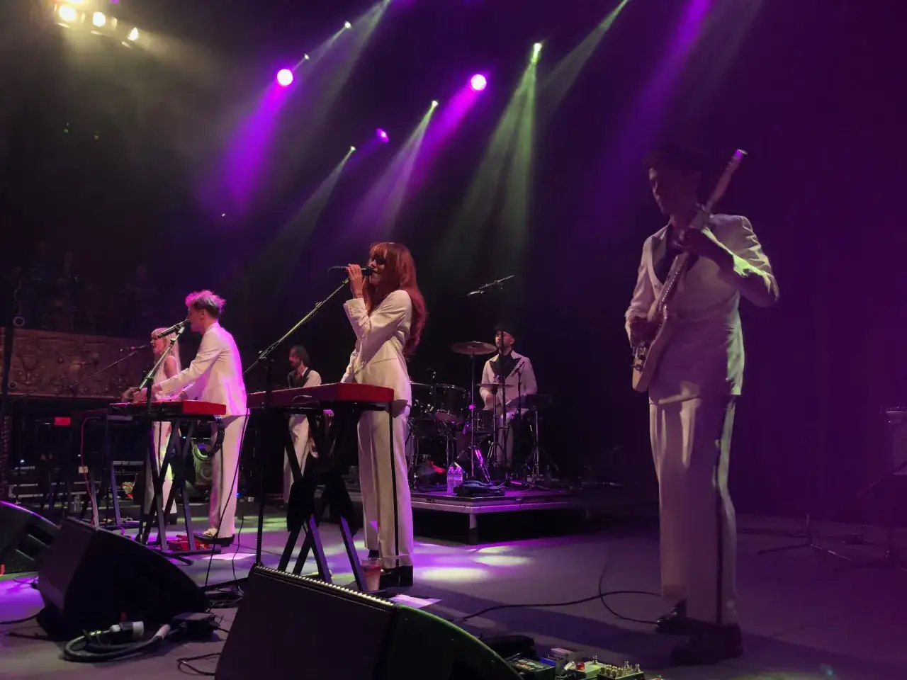 Zesp&oacute;ł la femme band w białych strojach na scenie, oświetlony fioletowymi i zielonymi światłami. Wokalistka śpiewa do mikrofonu, gitarzysta gra na gitarze, a perkusista siedzi za zestawem perkusyjnym.