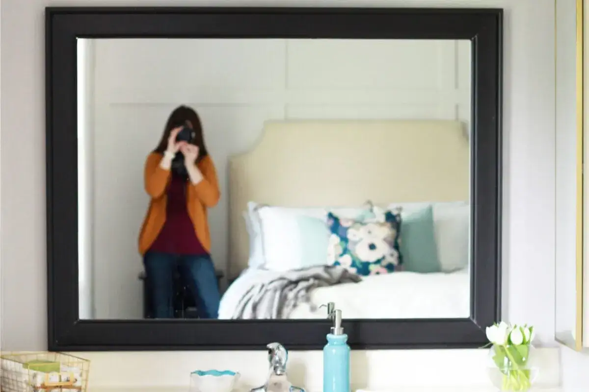 A person takes a selfie in a mirror with a DIY mirror frame. The reflection shows a bedroom with a bed and pillows.