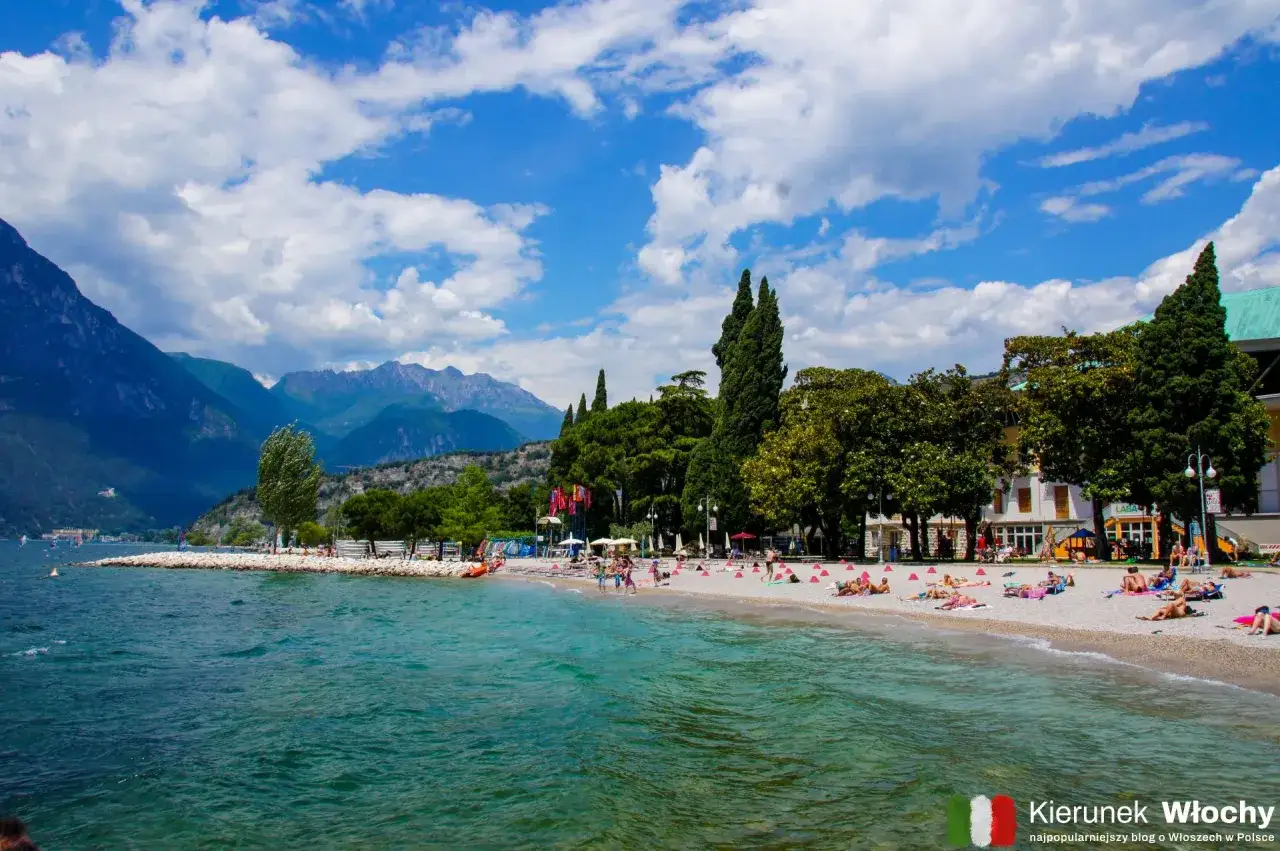 Jezioro Garda: plaża z ludźmi odpoczywającymi na kamieniach, w tle góry i drzewa. Idealne miejsce, by zobaczyć, jak cieszyć się włoskim słońcem.
