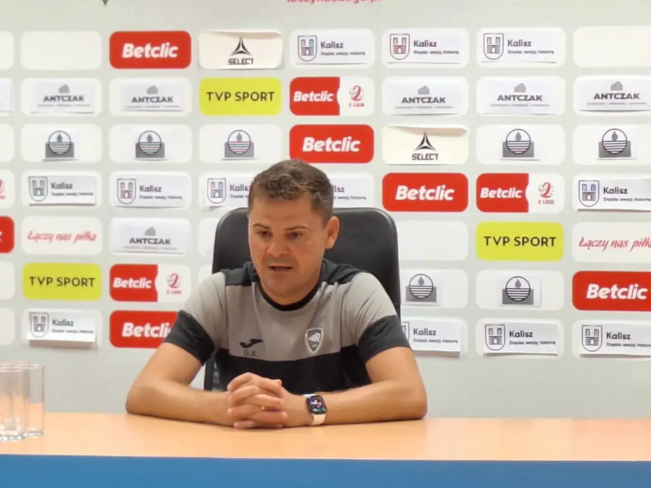 Maciej Kasprzak, trener, siedzi przy stole na tle ścianki z logotypami sponsorów, m.in. Betclic i Kalisz.