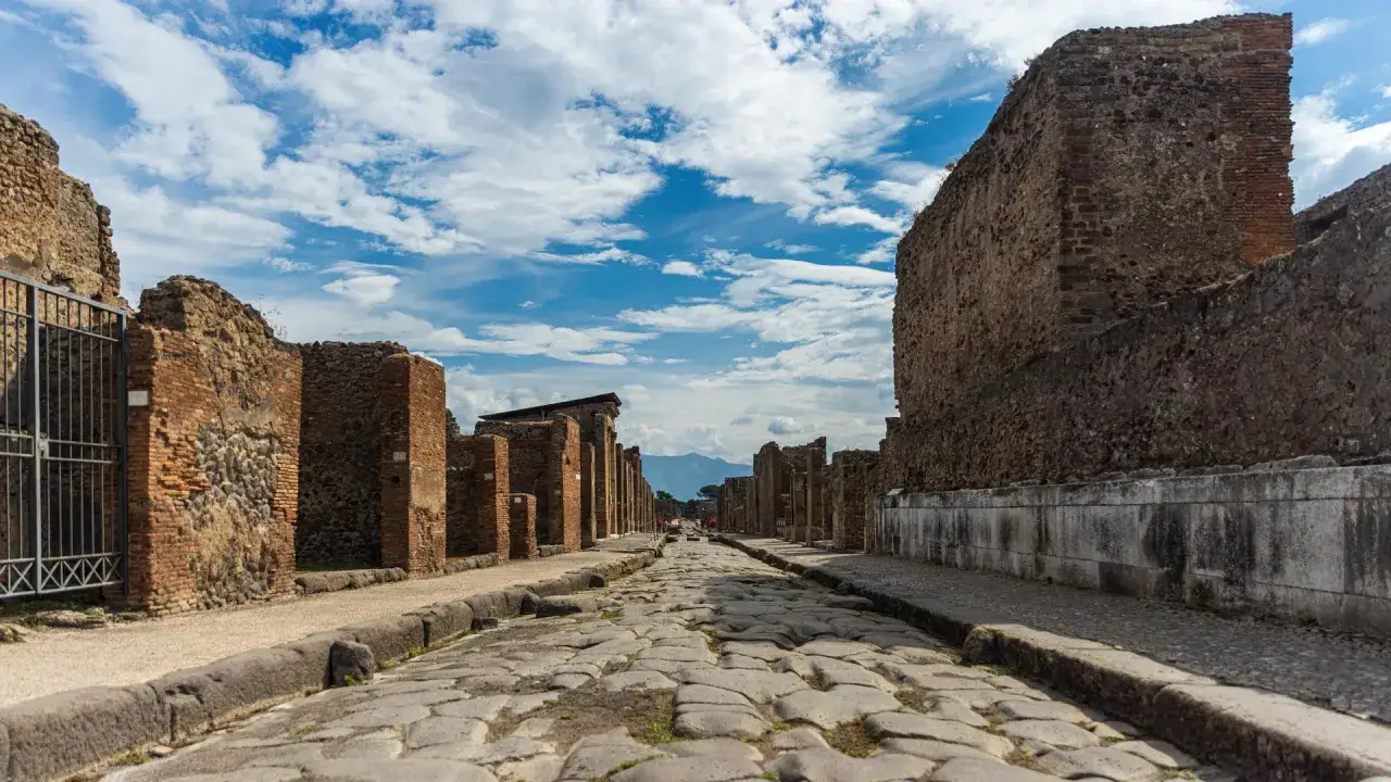 Antike Straße in Pompeji heute, gesäumt von Ruinen und unter blauem Himmel mit Wolken.