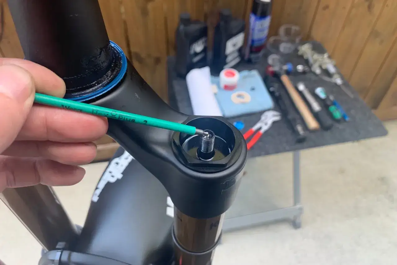 A hand uses a green hex wrench to adjust a bolt on a black bicycle fork, part of a Fox 34 service. Tools and lubricants are visible in the background.