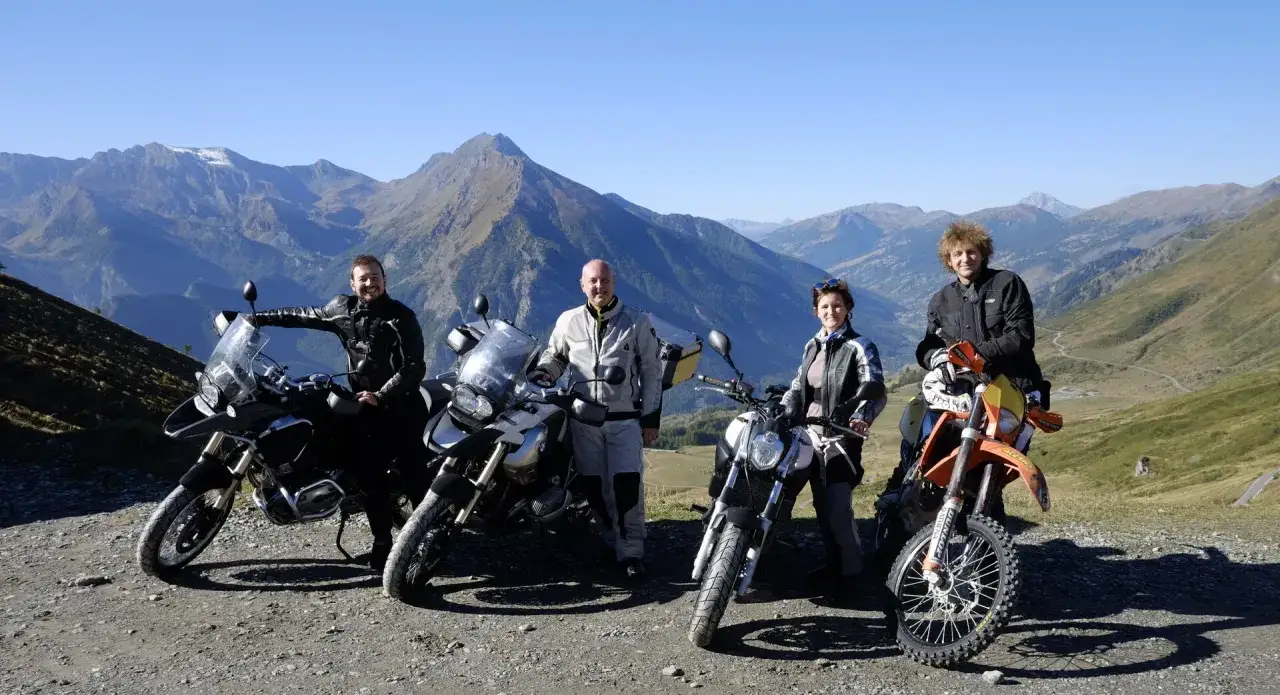 Quattro motociclisti posano con le loro moto su una strada panoramica, il Colle delle Finestre, con montagne imponenti sullo sfondo.